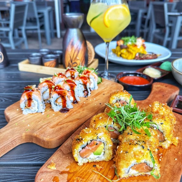 Two wooden boards with various sushi rolls, sauces, and a drink on a dark table.