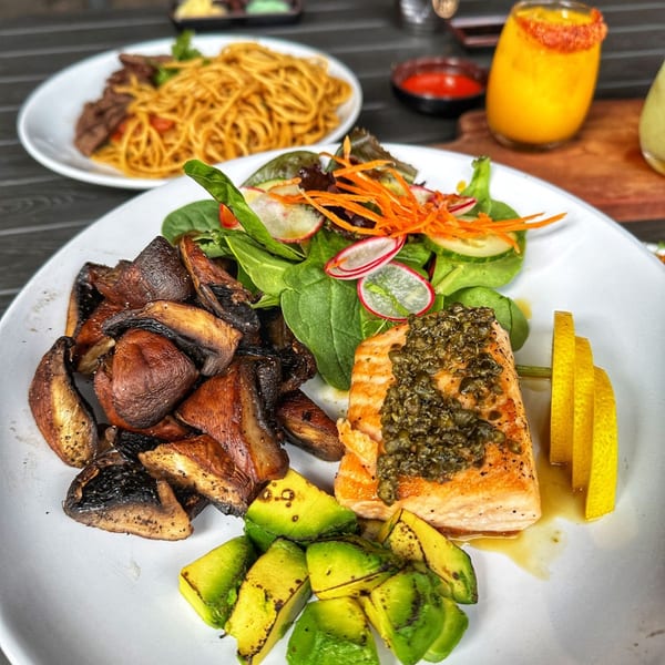 Grilled salmon with capers, roasted mushrooms, avocado, and a side salad, with noodles in the background.